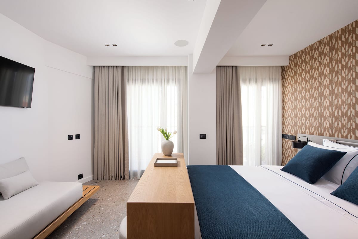 A modern hotel room with a bed, a sofa, a wooden side table, and a TV mounted on the wall.