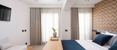 A modern hotel room with a bed, a sofa, a wooden side table, and a TV mounted on the wall.