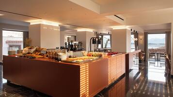 A modern hotel lobby with a central island featuring a buffet, a coffee station, and a seating area with a view of the city.