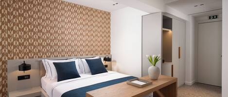 A modern hotel room with a neatly made bed, a wooden desk, a vase with flowers, and a patterned wall.
