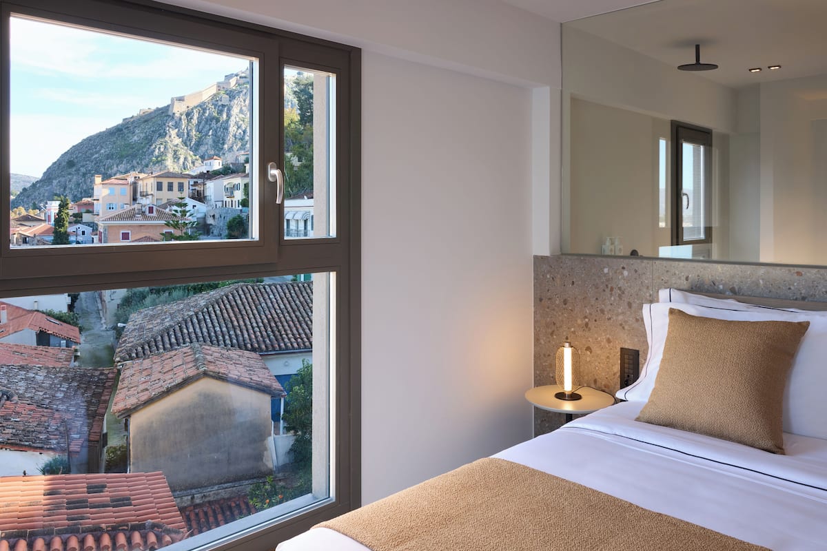 A bedroom with a large window, a bed with a beige blanket, a bedside table with a lamp, and a mirror above the bed.