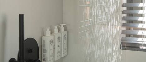 Chuveiro com porta de vidro, chuveiro manual e três frascos de shampoo na parede.