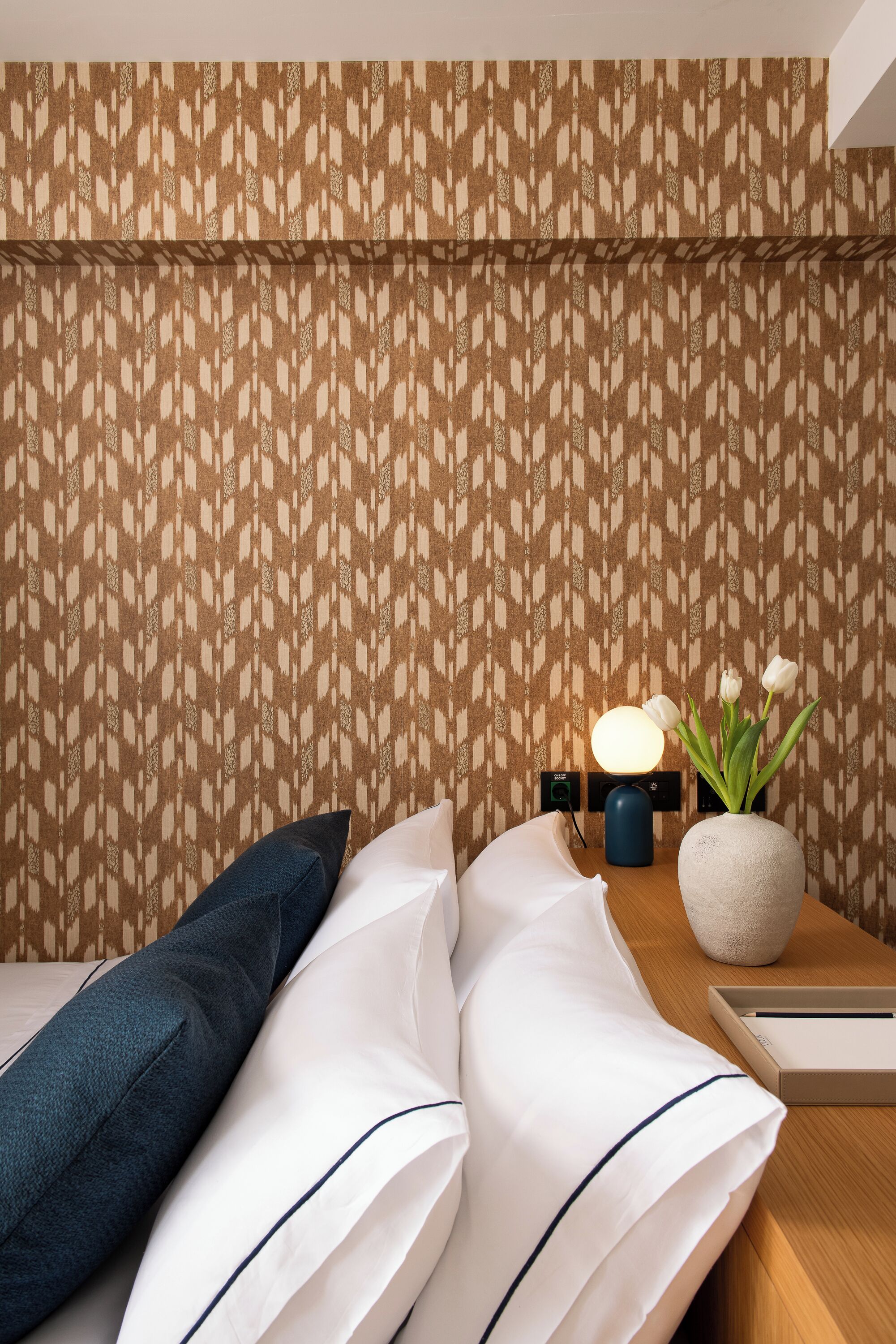 A neatly arranged hotel room with a patterned wall, a bedside table with a lamp, a vase with flowers, and a bed with pillows.