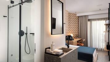 A modern hotel room with a glass-enclosed shower, a sink with a marble countertop, a bed with a blue bedspread, a wooden desk, and a large mirror.