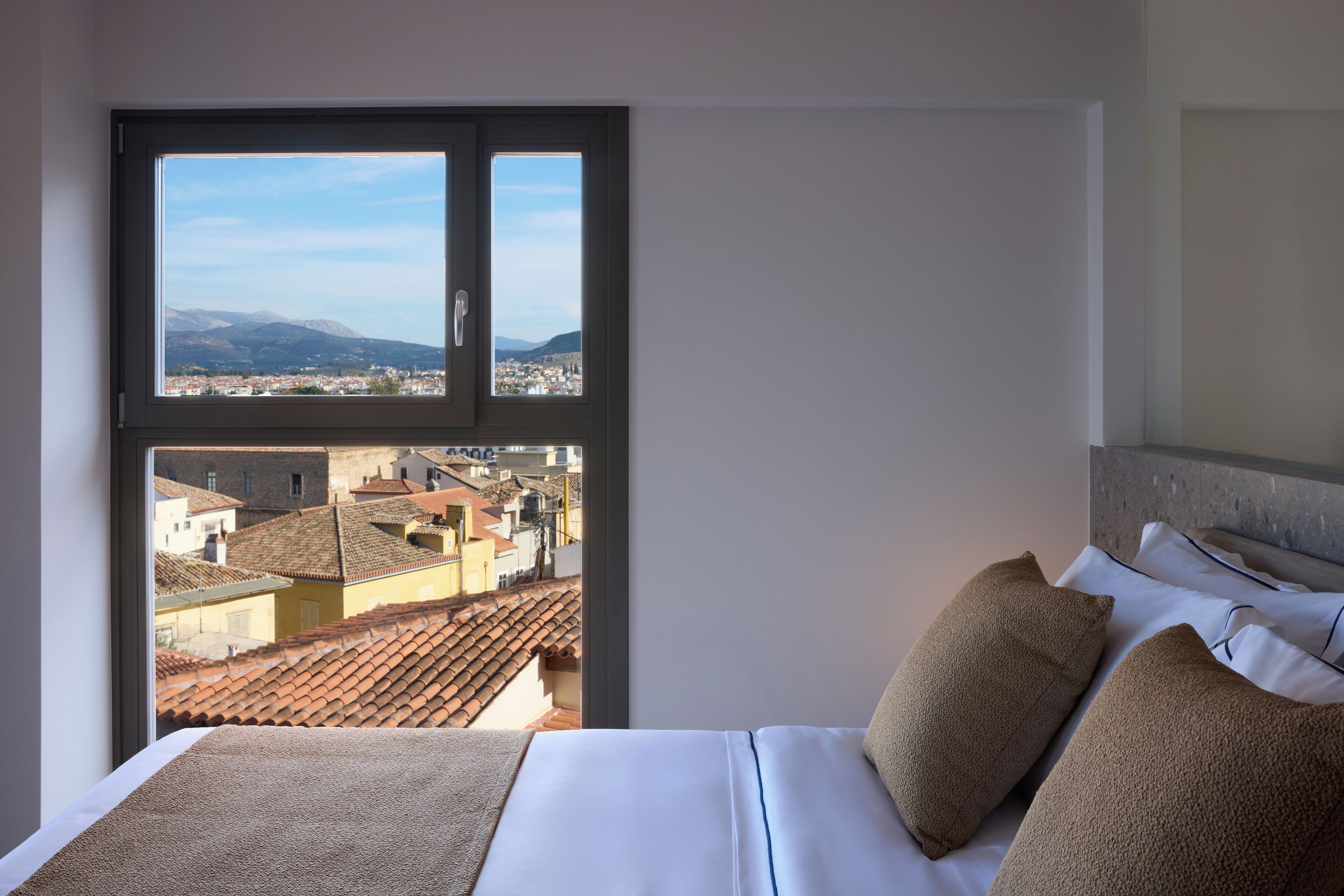 A hotel room with a large window offering a cityscape view, a neatly made bed, and a headboard with a textured design.