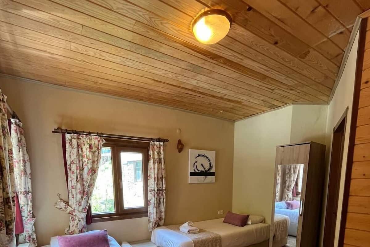A cozy room with two beds, a window with floral curtains, a wooden ceiling, and a small artwork on the wall.