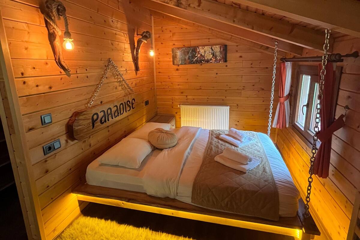 A wooden cabin bedroom with a bed, a fluffy rug, a wooden wall with a 'Paradise' sign, and a rustic decor including a skull and a lantern.