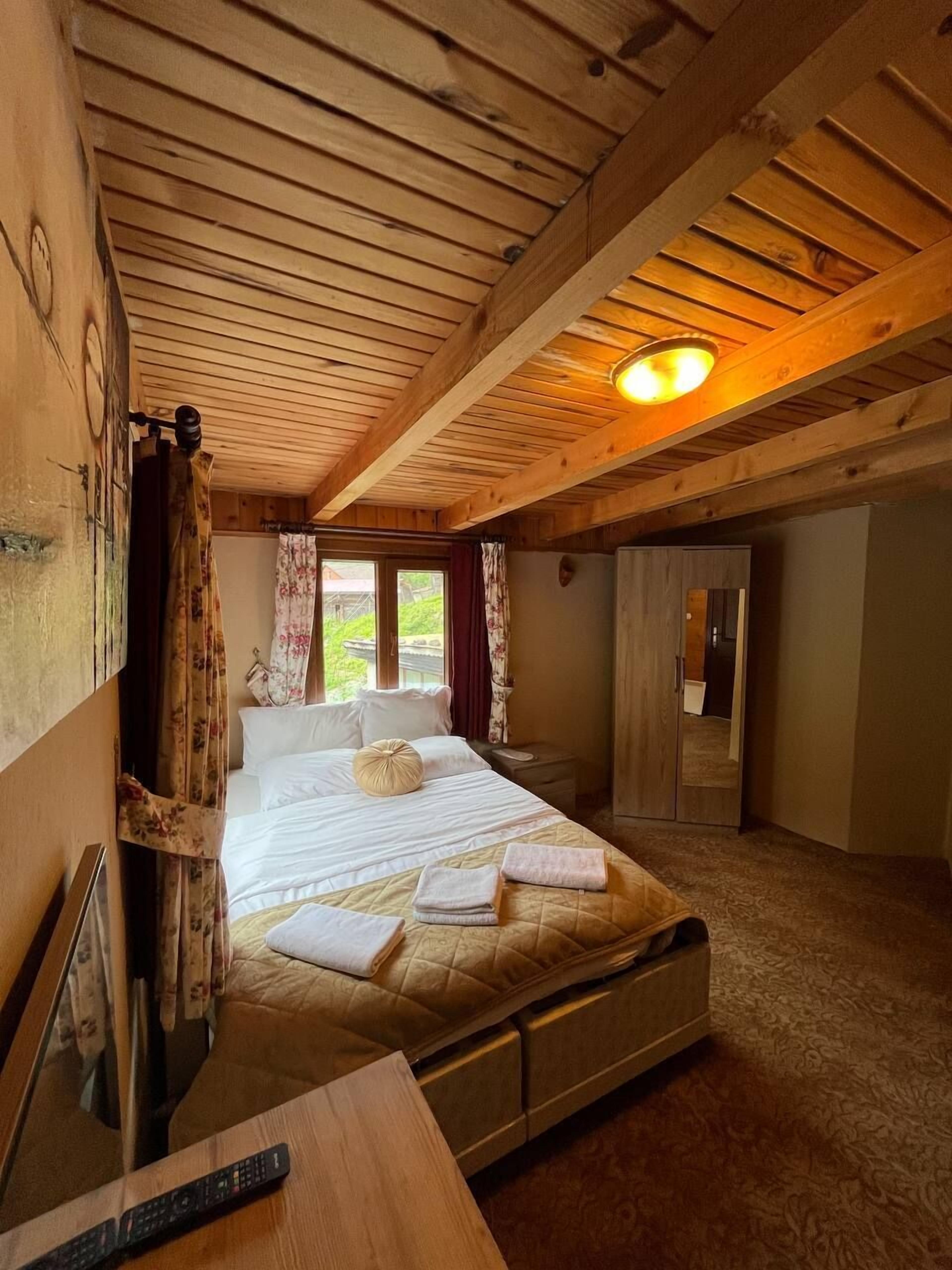 A cozy bedroom with a wooden ceiling, a large bed with white linens, a bedside table with a remote control, and a view of the outdoors through the window.