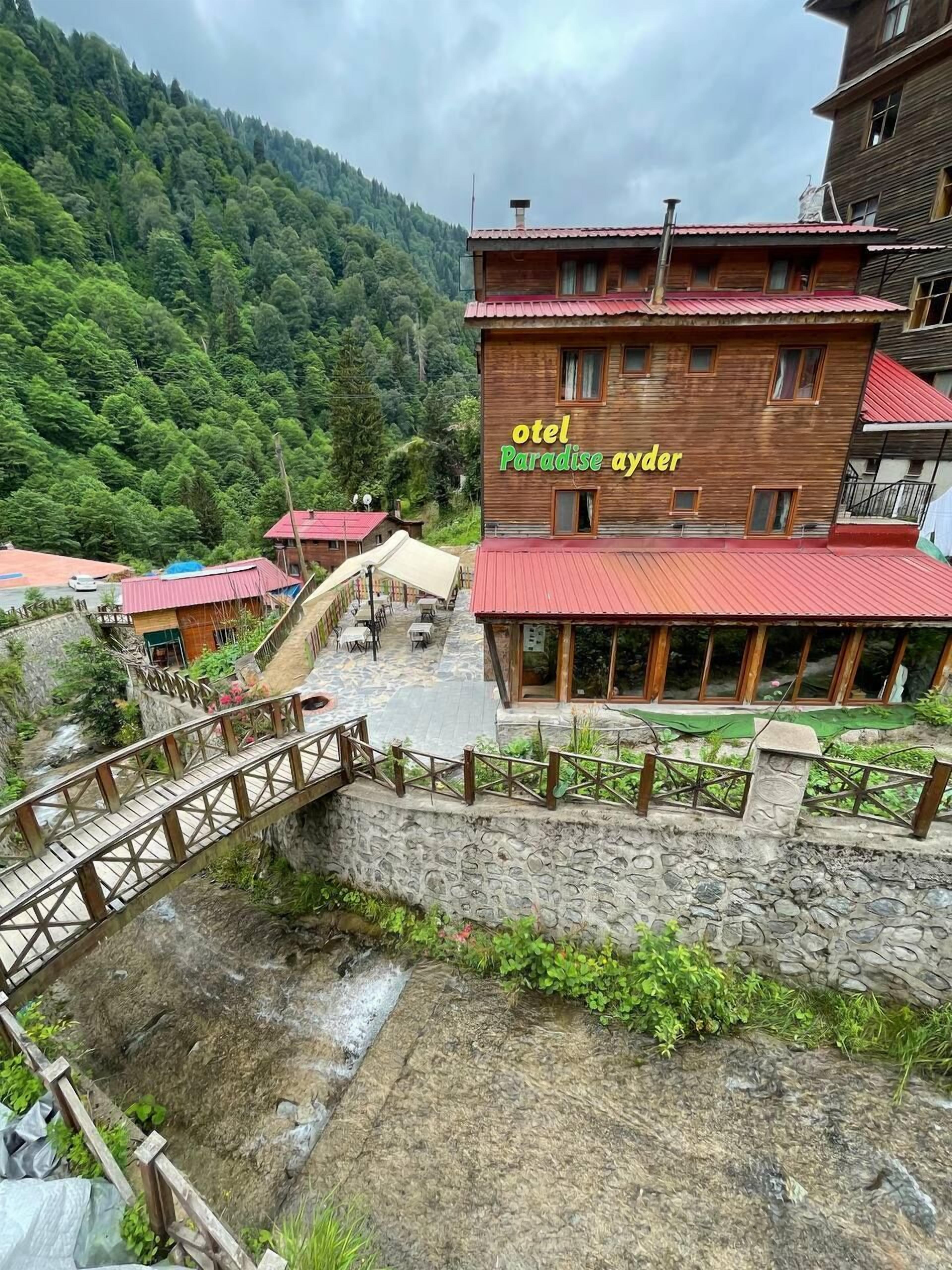 A wooden building with a red roof, labeled "otel Paradise ayder," surrounded by lush greenery and a stone bridge.