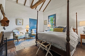 Un lit à baldaquin avec un cadre en bois, une porte bleue, une petite table ronde avec une lampe et une chaise avec un coussin à carreaux.