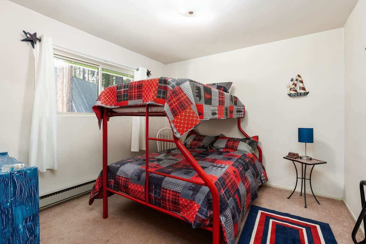 Ein Stockbett mit rotem Metallgestell und einer karierten Decke. Ein kleiner Tisch mit Lampe, eine Stern-Dekoration an der Wand und ein Fenster mit Vorhängen.