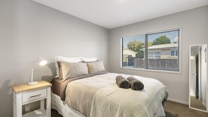 A bedroom with a bed, bedside table, lamp, and a window with blinds. - Sandy Feet Retreat - Papamoa (Papamoa)