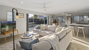 A modern living room with a large sofa, a coffee table, and a TV. There is a balcony with a table and chairs. - Sandy Feet Retreat - Papamoa (Papamoa)