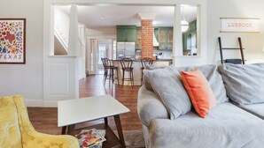A living room with a grey sofa, a yellow armchair, and a white table. There is a kitchen in the background with green cabinets and a brick column.