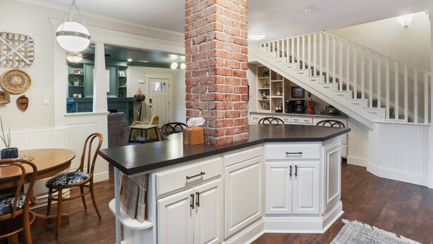 A kitchen with white cabinetry, a brick column, a dining area with a wooden table and chairs, and a staircase with white railings.