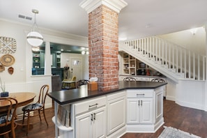 A kitchen with white cabinetry, a brick column, a dining area with a wooden table and chairs, and a staircase with white railings. - Brick Road Retreat - 3BR/2BA - Walk to Tech (Lubbock)