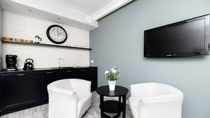 Ett modernt kök med vit tegelvägg bakom arbetsbänken, svarta skåp, en vit soffa, ett litet svart sidobord med en vas och en väggmonterad platt-TV.