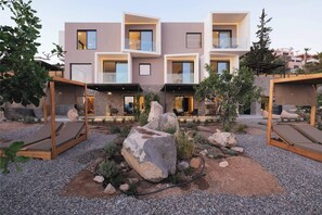 Un edificio moderno de varios pisos con balcones, un jardín ajardinado con rocas y plantas, y zonas de estar al aire libre.
