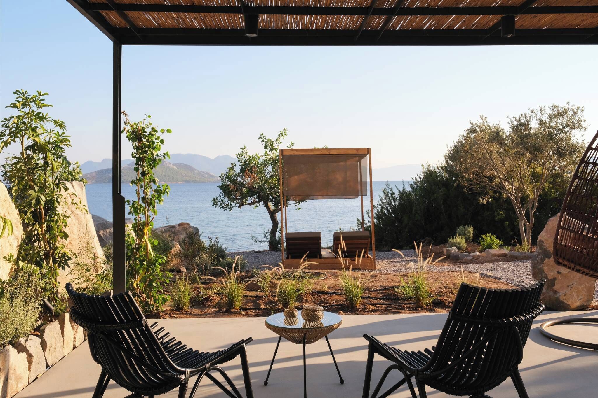A patio with two black wicker chairs, a small round table, and a wooden swing chair overlooking a body of water with mountains in the background.
