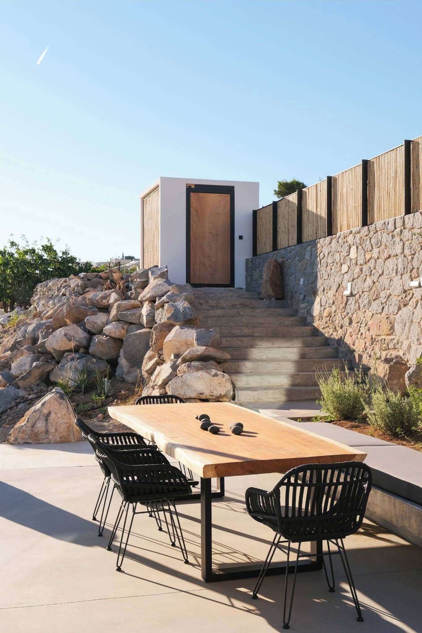 Une terrasse avec une table en bois et des chaises noires, un mur en pierre et une porte en bois donnant accès à un bâtiment.