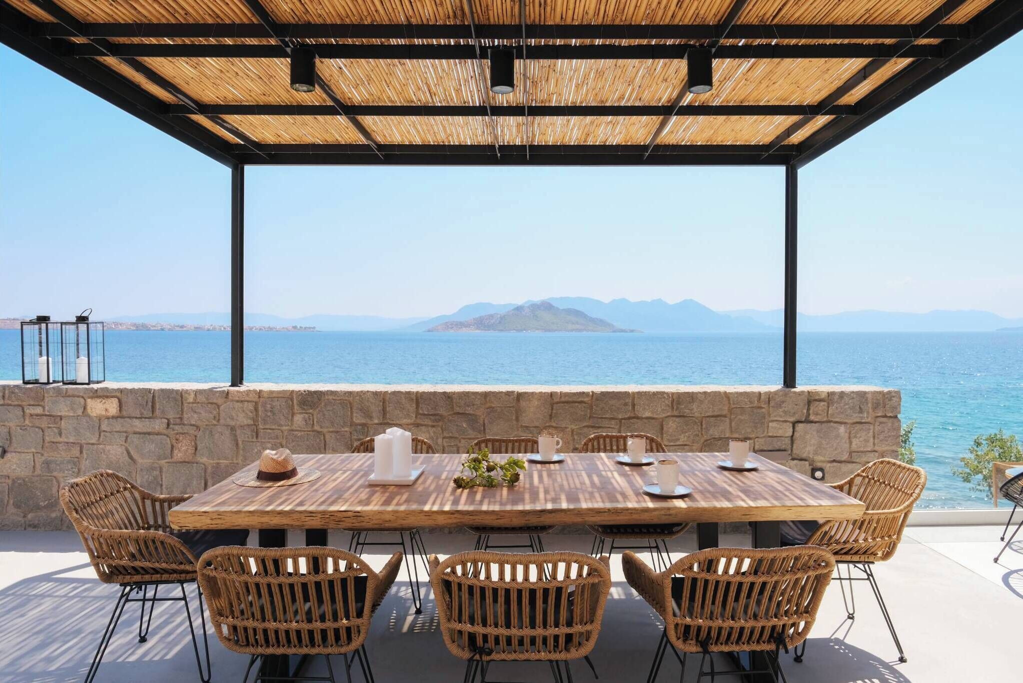 A wooden table with wicker chairs, set against a stone wall with a view of the sea and mountains.