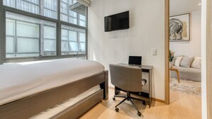 Quarto com cama, escrivaninha, laptop, televisão e uma grande janela.