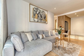 A modern living room with a grey sectional sofa, a framed elephant artwork, a small round table with a vase, and a wooden dining area in the background.