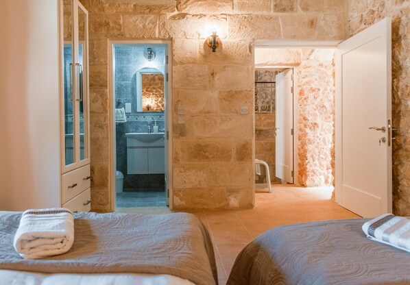A bedroom with two beds, a bathroom with a stone wall, and a door leading to another room. - Villa Oliva Sleeps 8 With Gym (Xaghra)