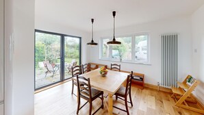 Un comedor con una mesa de madera, sillas y un bol de fruta. Hay dos ventanas y un radiador en la pared.