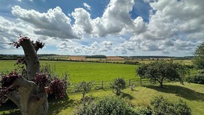 Paisaje rural con cielo despejado, campos verdes y una construcción de piedra con follaje rojo.