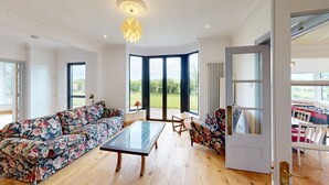 Amplia sala de estar con un sofá floral, una mesa de centro de vidrio y vistas al exterior a través de amplias ventanas.