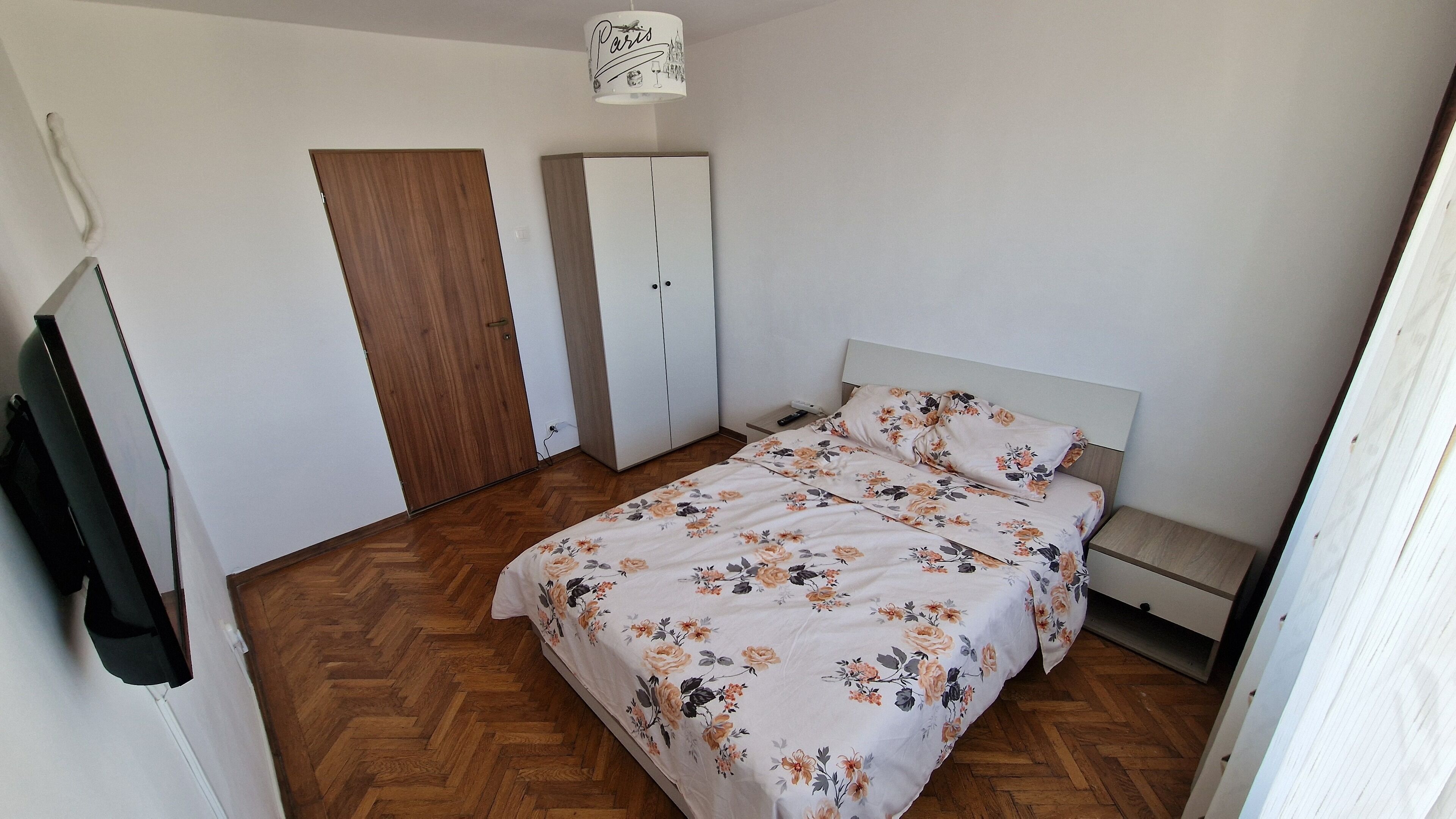 Een slaapkamer met een houten visgraatvloer, een bed met bloemenbeddengoed, een garderobe en een televisie.