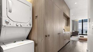Une buanderie moderne équipée d’un lave-linge et d’un sèche-linge à chargement frontal, de meubles en bois et offrant un aperçu d’une chambre avec un lit et une fenêtre.