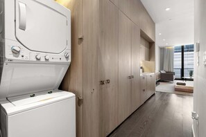 Een moderne wasruimte met een voorlader wasmachine en droger, houten kasten, en een blik op een slaapkamer met een bed en een raam.