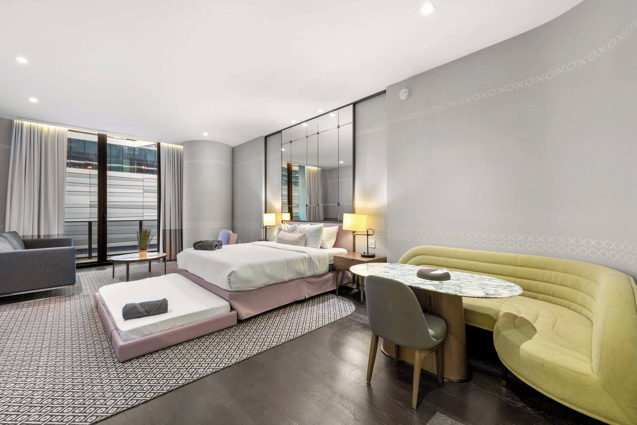 A modern hotel room with a bed, a sofa, a dining table, and a large mirror.
