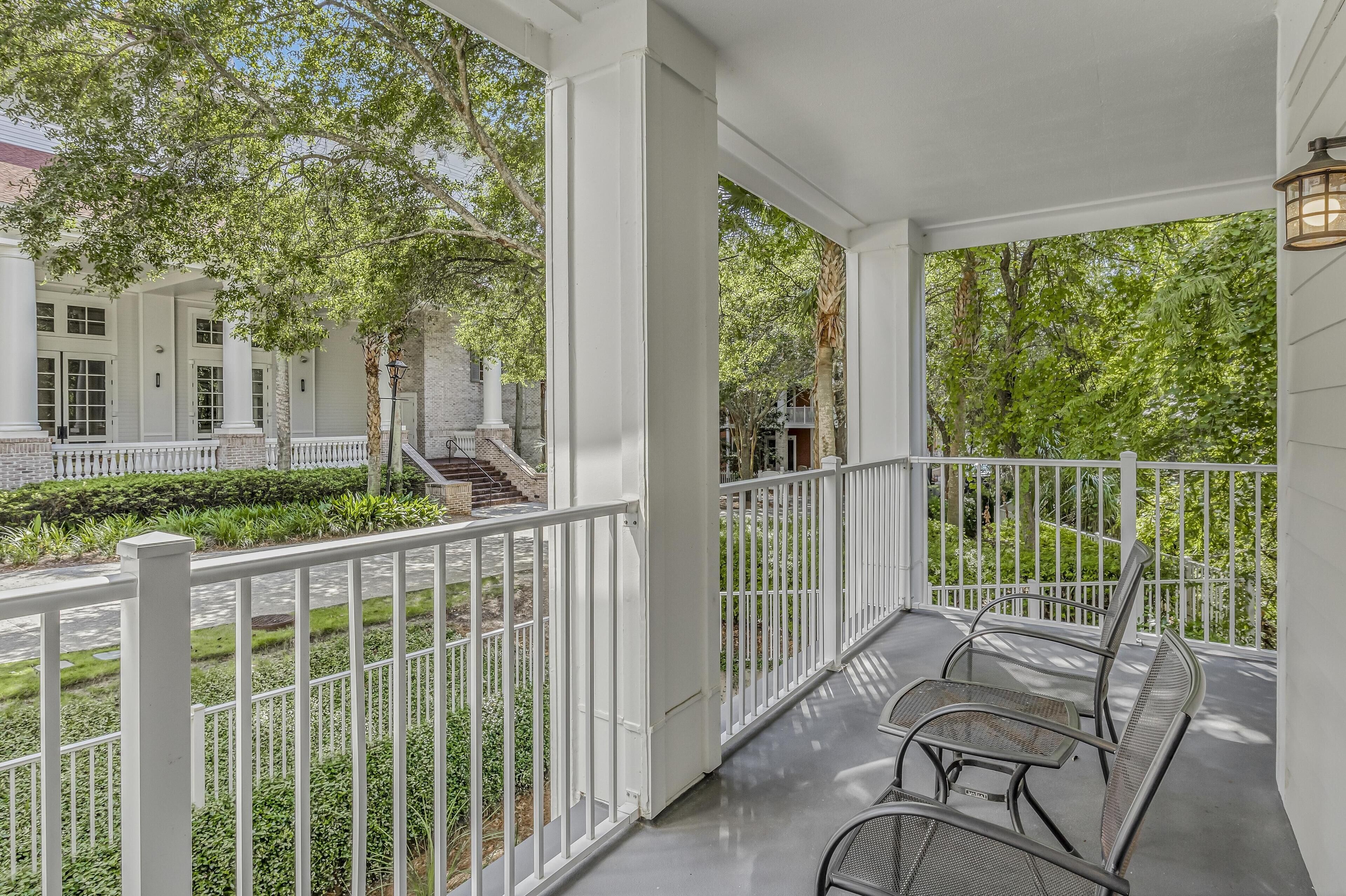 A covered porch with white railings, a table, and chairs, overlooking a landscaped garden and a building with columns.
