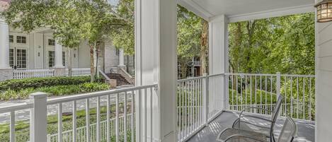 A covered porch with white railings, a table, and chairs, overlooking a landscaped garden and a building with columns.