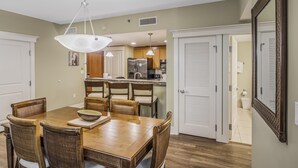 A dining area with a wooden table and chairs, a hanging light fixture, and a kitchen in the background. - Grand Comfort: 2 BR w/ Golf Cart & Bay Views at Sandestin Golf and Beach Resort! (Miramar Beach)