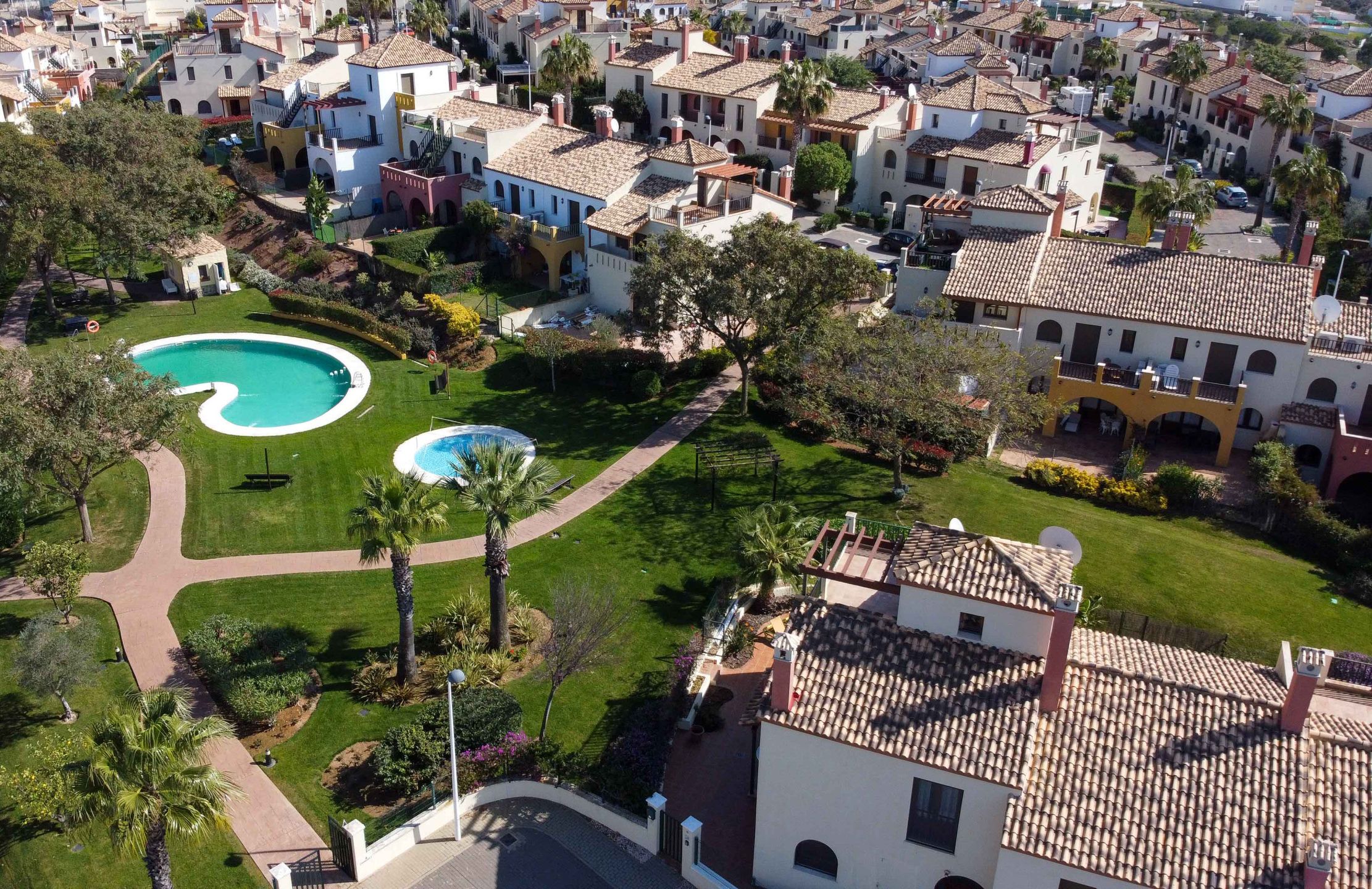 An aerial view of a residential area with two swimming pools surrounded by well-maintained lawns and palm trees.