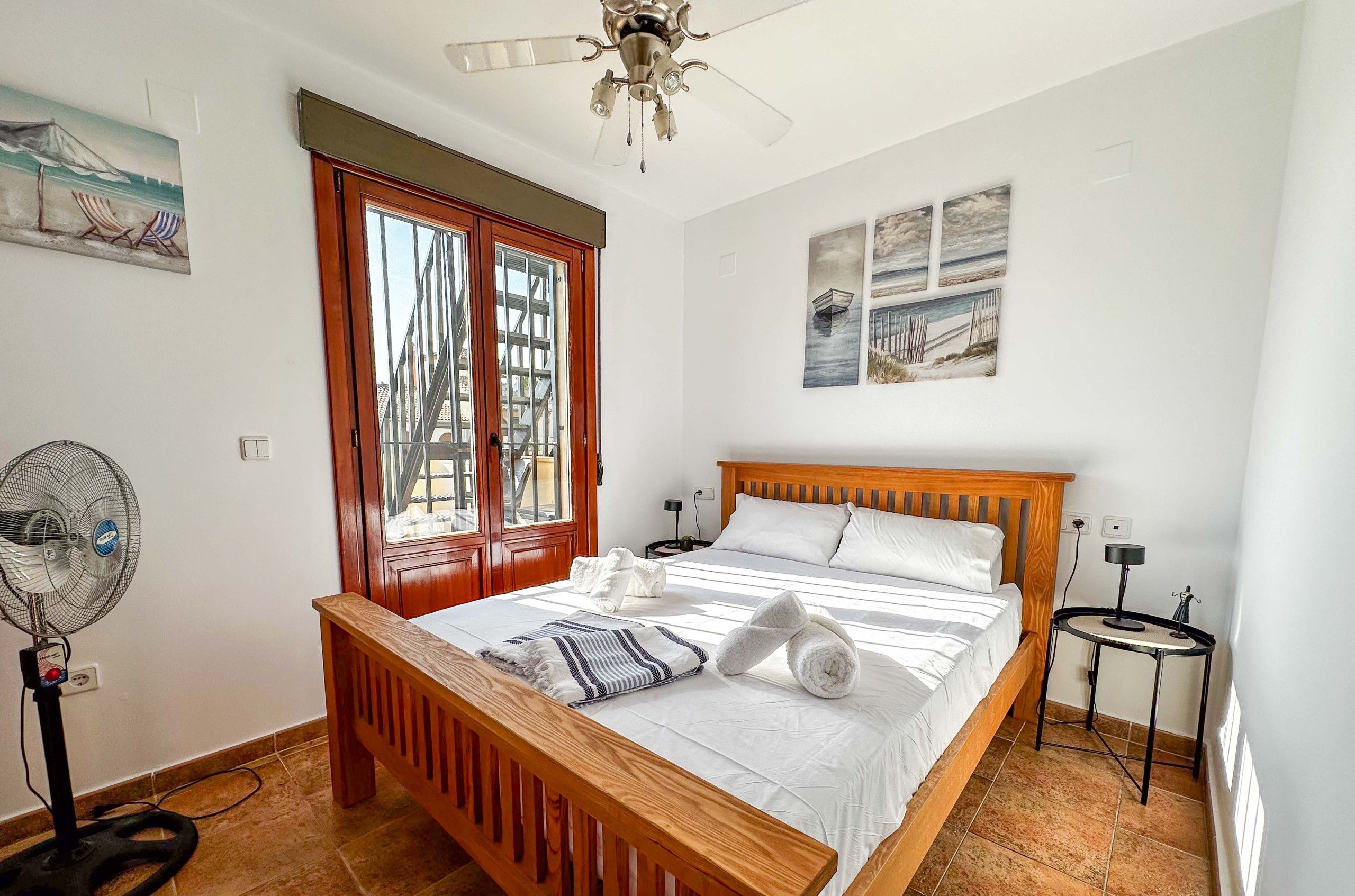 A bedroom with a wooden bed, a standing fan, and a view of a building through a window.