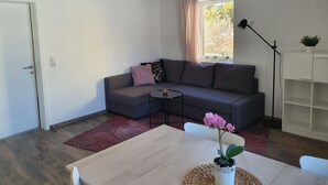 Una sala de estar moderna con un sofá gris, una pequeña mesa redonda, una mesa de centro de madera, un tapete rojo, una ventana con cortinas, una lámpara de pie y un estante blanco.
