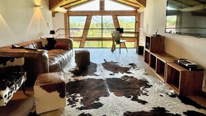Quarto com teto alto de madeira, uma grande janela, sofá de couro, tapete de couro bovino e TV fixada na parede.