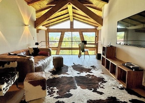 A room with a high wooden ceiling, a large window, a leather sofa, a cowhide rug, and a mounted TV.