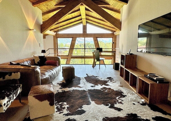 A room with a high wooden ceiling, a large window, a leather sofa, a cowhide rug, and a mounted TV.
