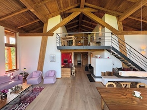 A spacious loft-style living area with a high ceiling, wooden beams, a mezzanine level, and a staircase.