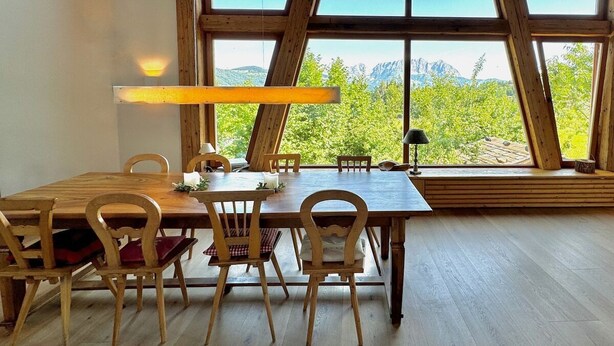 Uma sala de jantar com mesa e cadeiras de madeira, janelas amplas com vista para as montanhas e estrutura de teto em madeira.