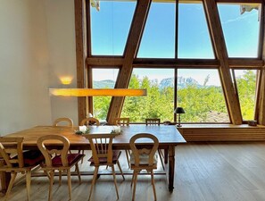 A dining area with a wooden table and chairs, large windows with a mountain view, and a wooden ceiling structure.