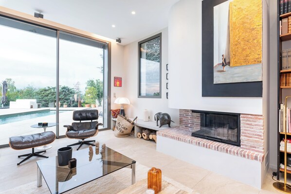 A modern living room with a fireplace, a glass coffee table, leather chairs, and a view of a swimming pool.
