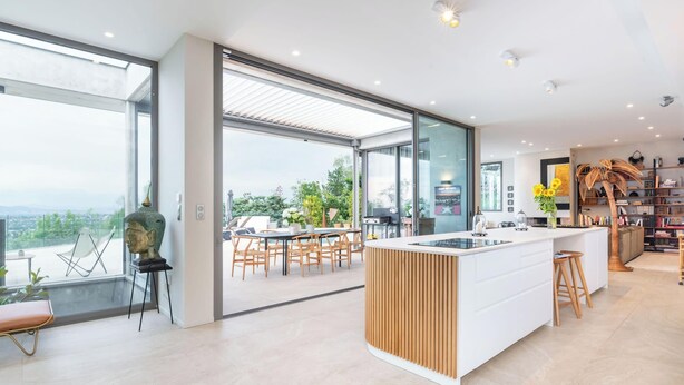 A modern kitchen with a large island, a dining area with wooden chairs, and a view of the city through large windows. - Futuriste - Exceptional residence with panoramic view (Saint-Didier-au-Mont-d'Or)