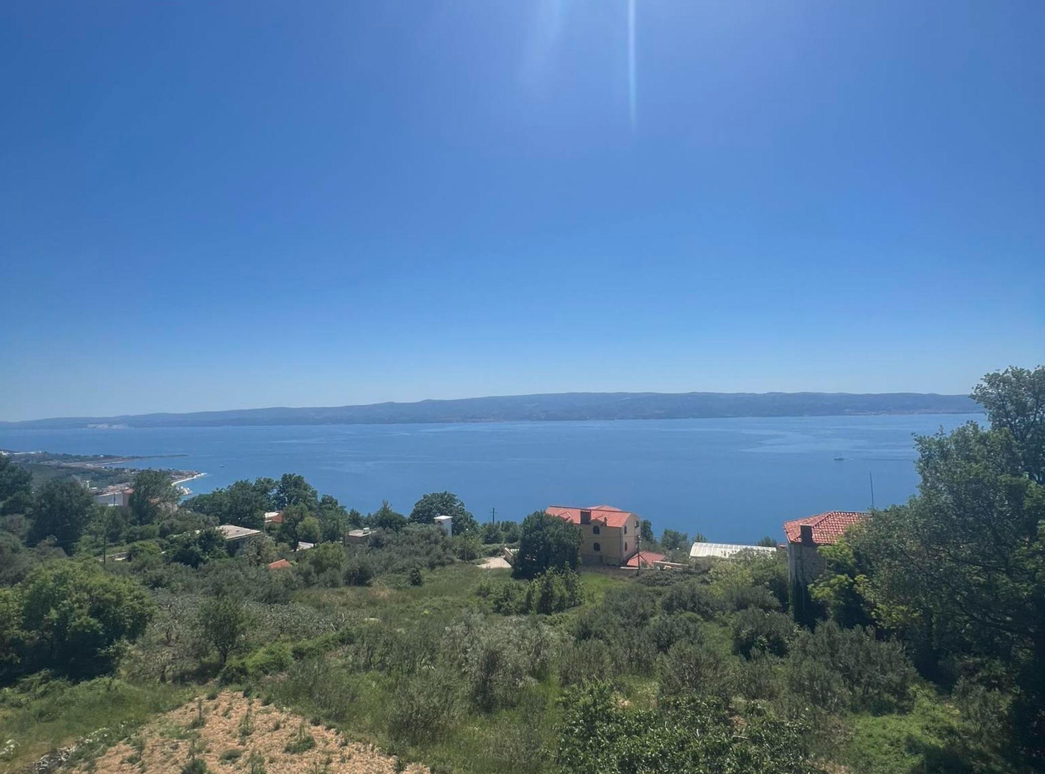 A coastal landscape with a clear view of the sea, a few buildings, and dense greenery.